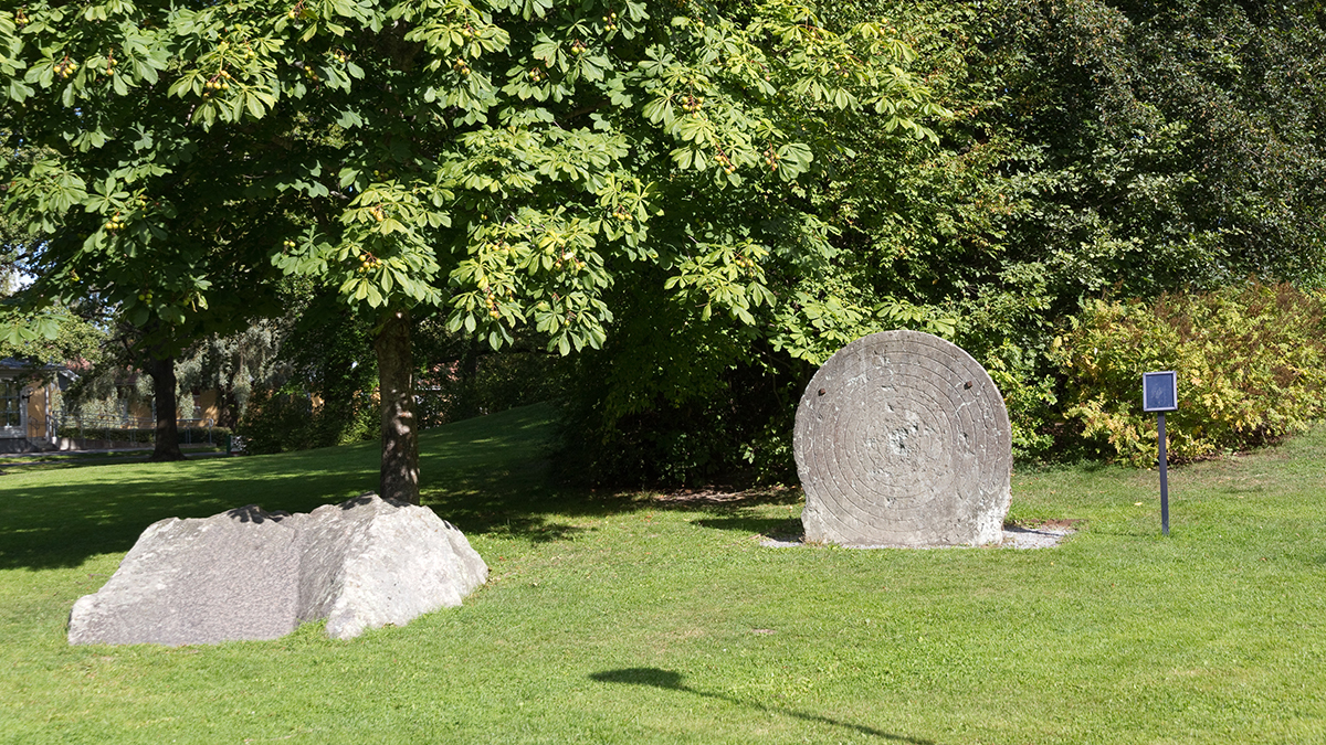 Skytteparken. Den runda skyttetavlan i sten står i mitten på bilden. Till vänster syns en stor grå stenbumling och i bakgrunden ett fullvuxet kastanjeträd. Till höger om måltavlan syns en blå liten skylt på en stolpe.