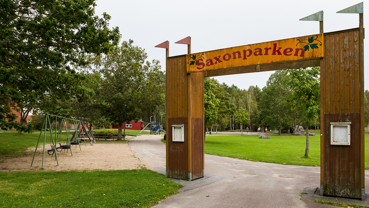Entrén till Saxons park med en portal i trä med fyra träflaggor och en skylt där det står Saxons park. Till vänster syns en rad med lövträd och en gungställning med sex gungor.