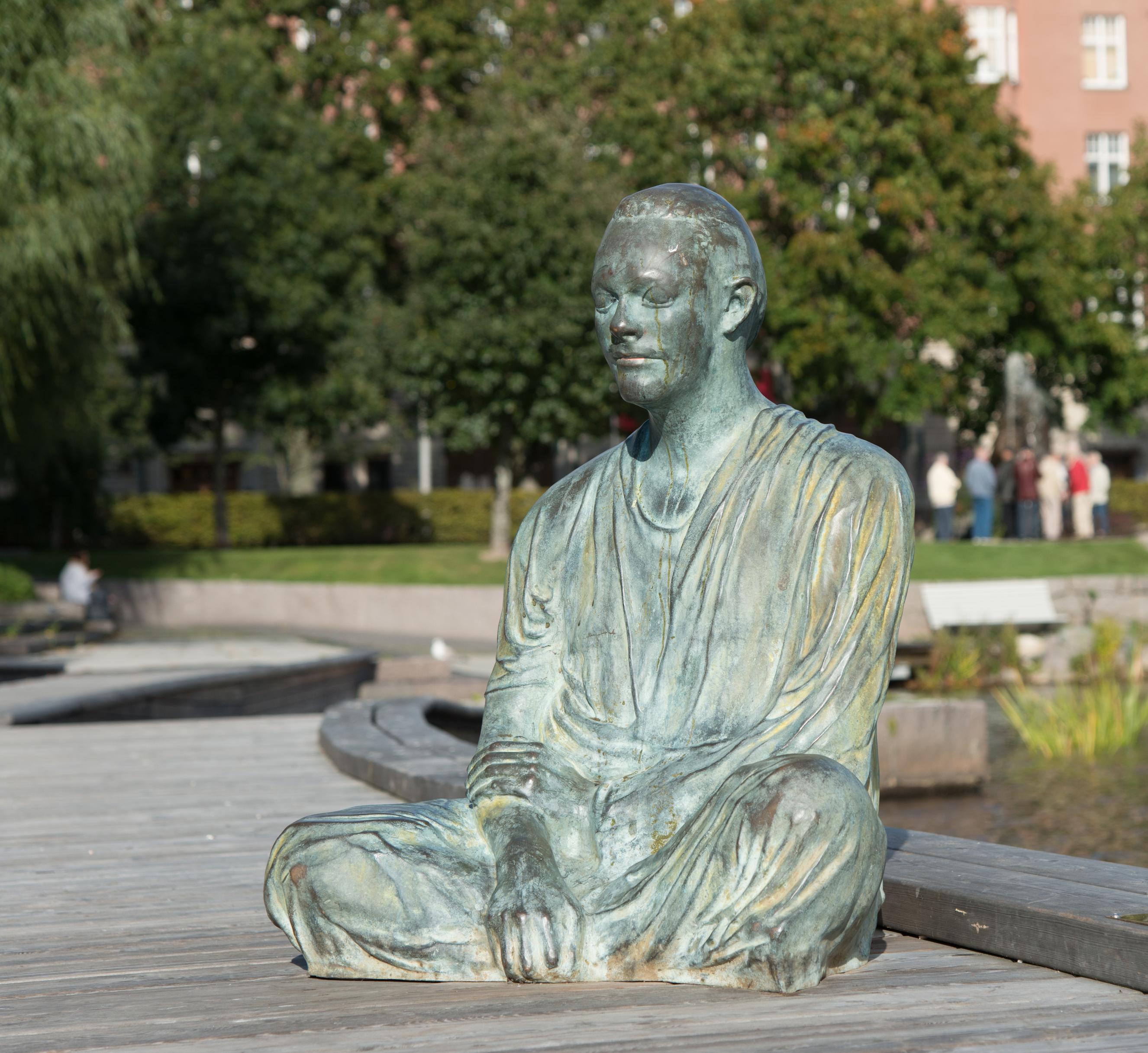 Bronsskulptur föreställande en man sitter i skräddarställning på soldäcket intill Svartån.