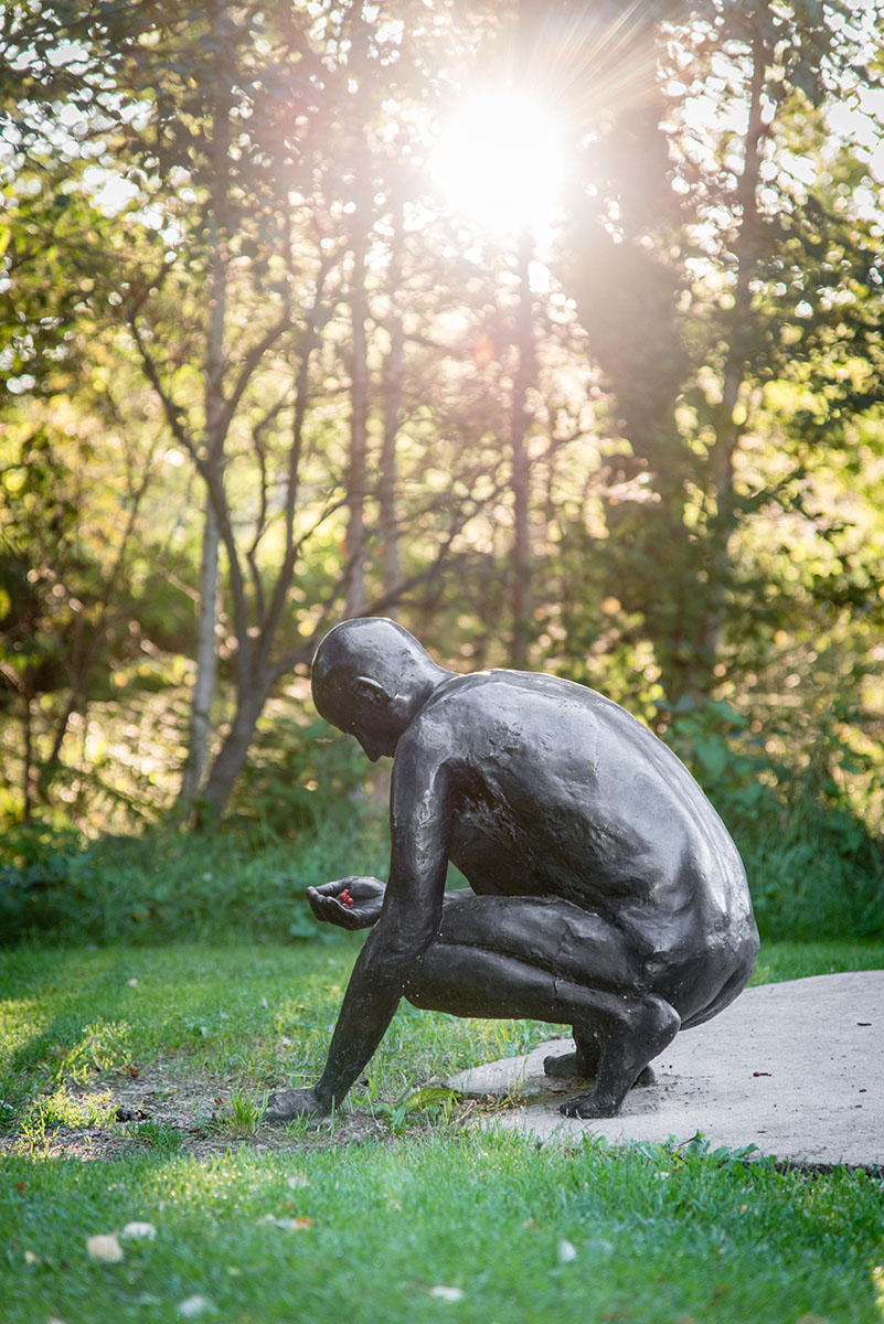 Skulptur av en man i betong och brons som är knäböjande och har plockat upp något från marken.
