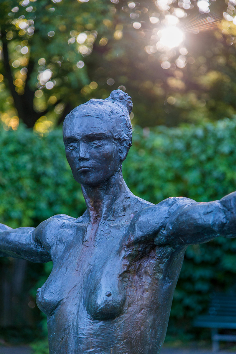 Tvärs över en liten damm i Stadsparken balanserar en gjuten skulptur av en kvinna med utsträckta armar på en smal ribba.