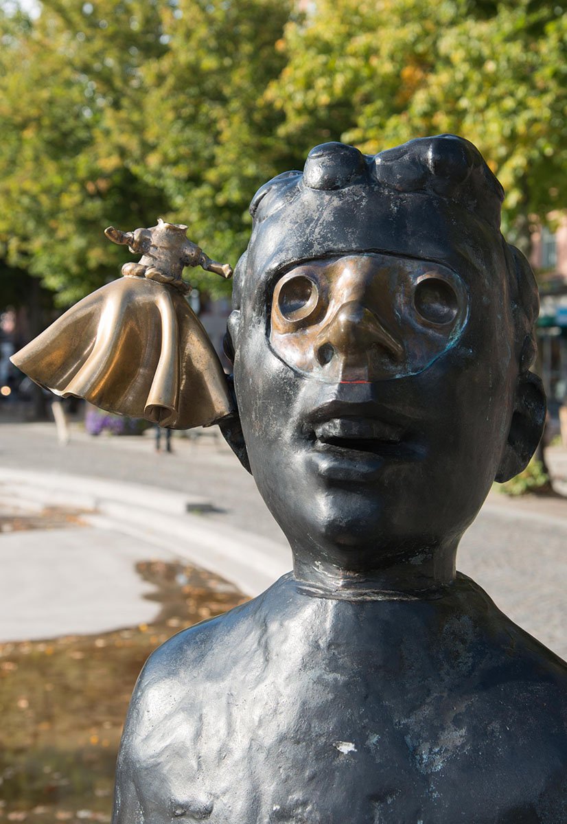 Mörk bronsfigur föreställande en pojke med djupa ögonhålor och öppen mun. Ansiktet ser ut att vara täckt av en mask och på sidan svävar en kvinna utan huvud.
