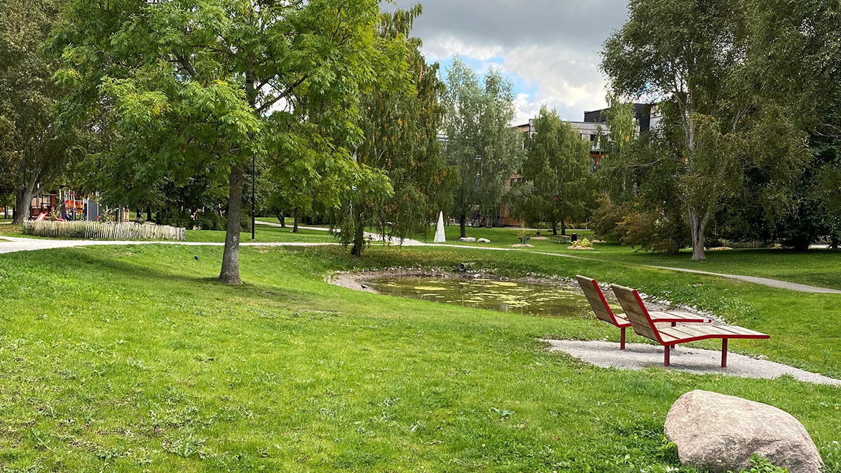 Grön gräsmatta, sittplatser vid en damm, i bakgrunden syns träd.