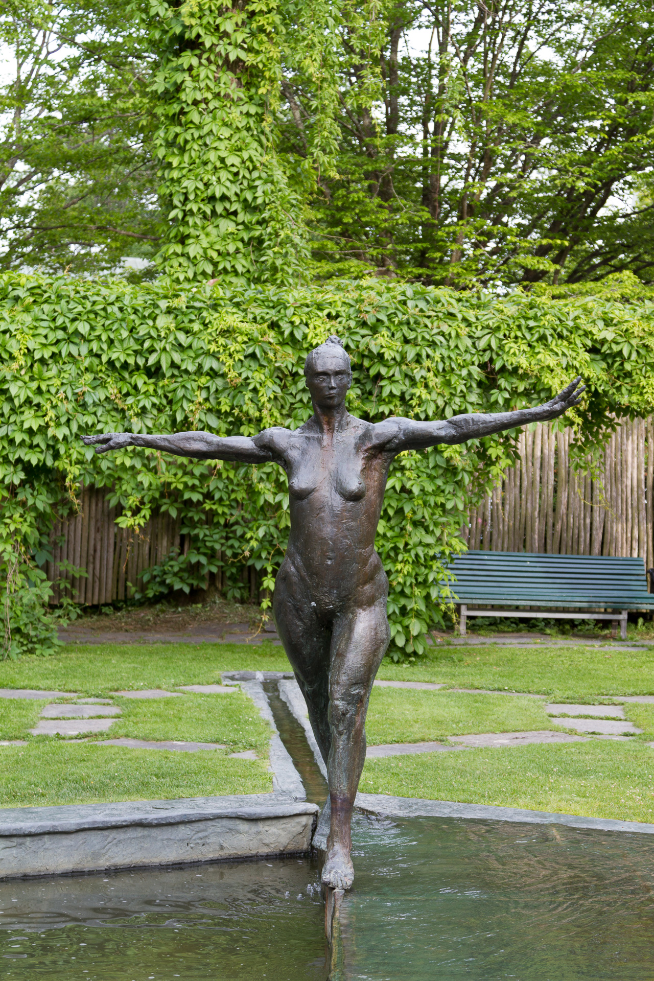 Tvärs över en liten damm i Stadsparken balanserar en gjuten skulptur av en kvinna med utsträckta armar på en smal ribba.