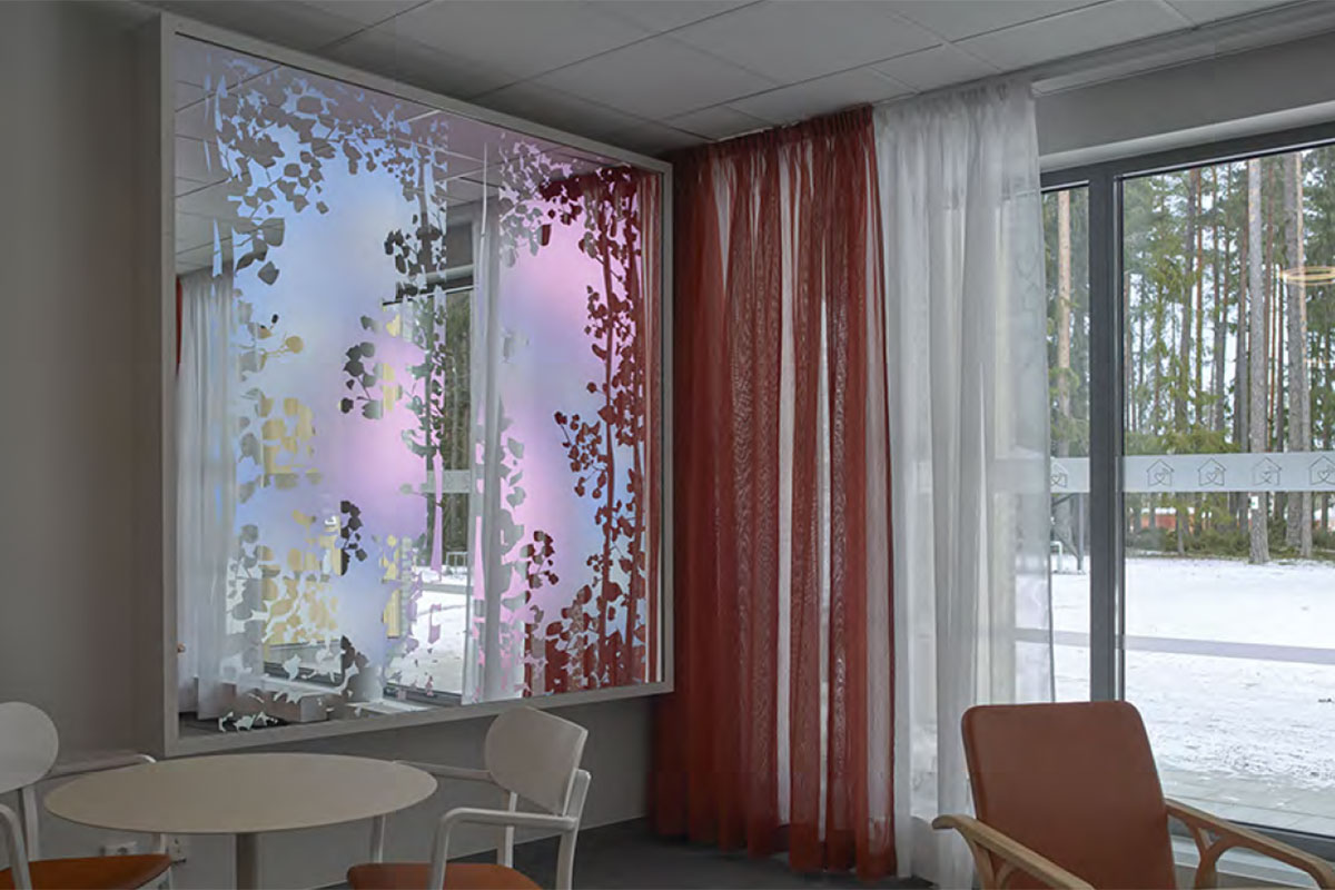 På väggen vid sidan om ett heltäckande glasfönster hänger en vit ram där lövverkssilhuetter ramar in ett skiftande ljusspel