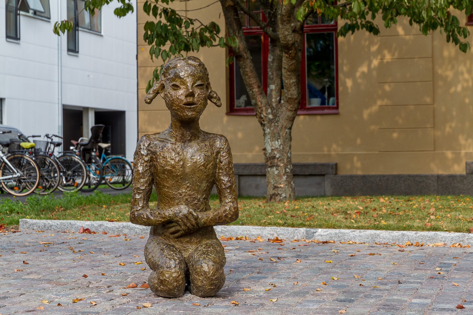 Staty i brons av naken flicka med flätor sittande på knä.